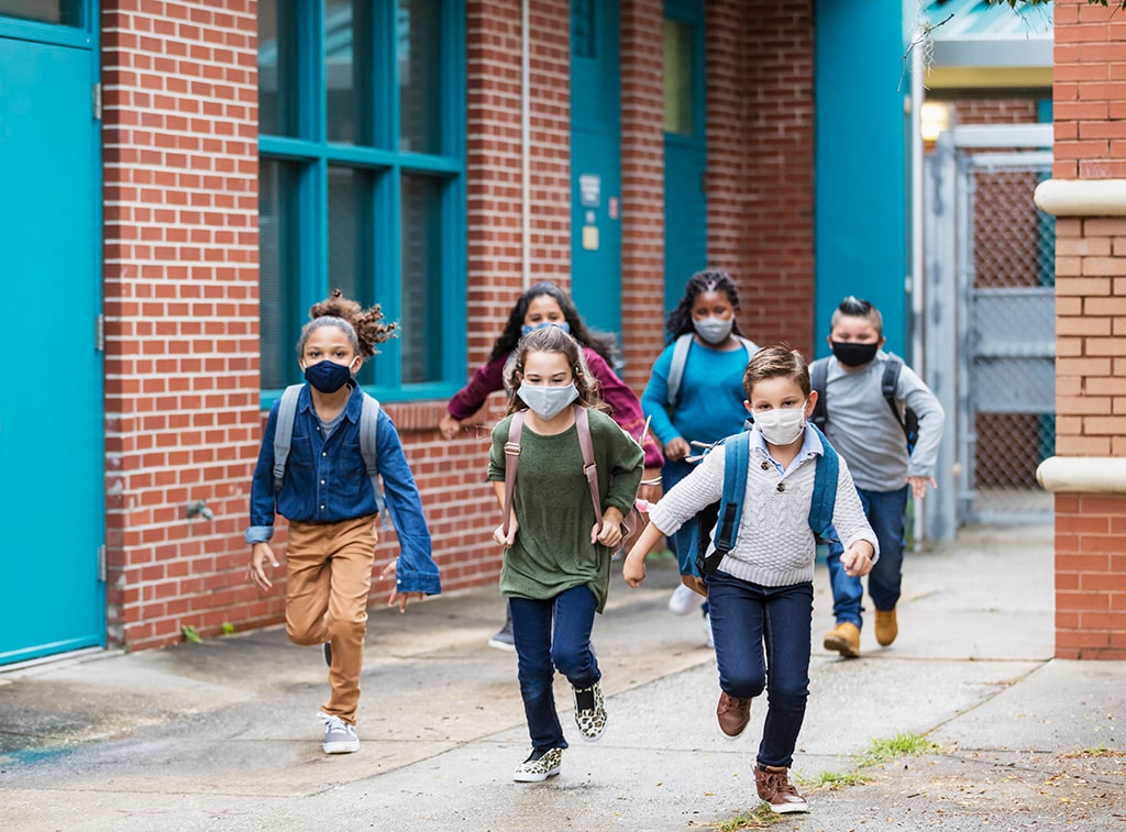 six children wearing masks and backpacks run in a schoolyard