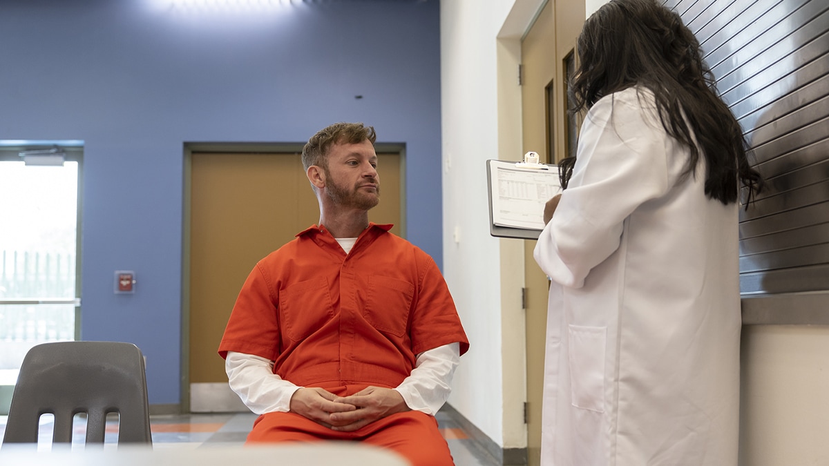 A man in an orange jumpsuit listens to a person in a white coat with a clipboard.