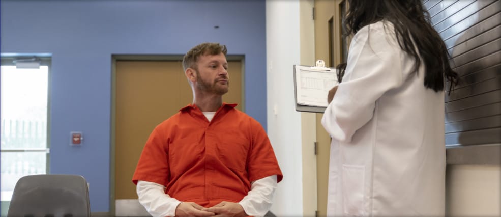 A man in an orange jumpsuit listens to a person in a white coat with a clipboard