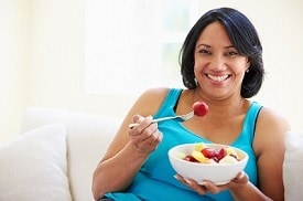 Una mujer con sobrepeso sentada en un sofá comiendo un tazón de fruta fresca