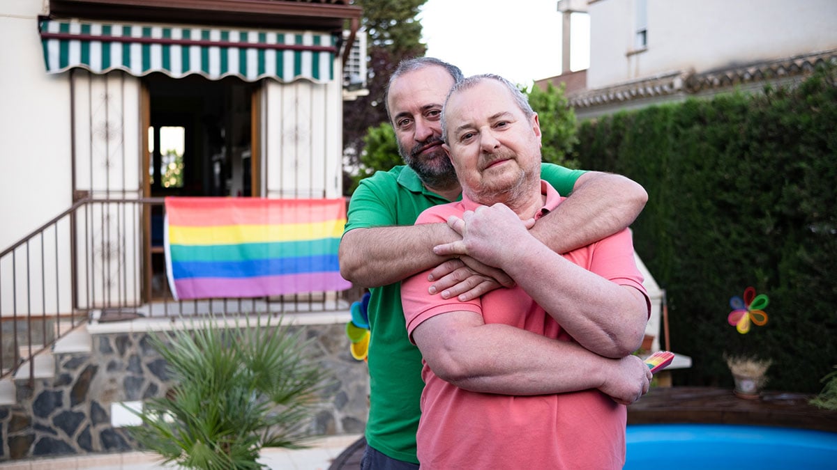 LGBTQ couple embracing in their back yard with a LGBTQ flag in the background.