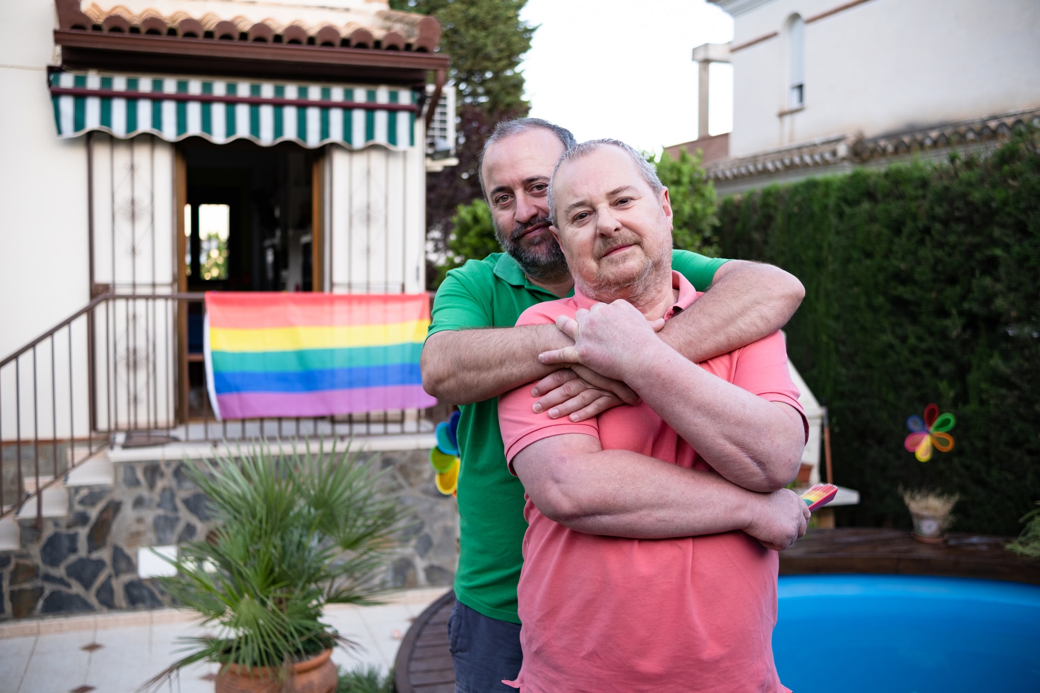 LGBTQ couple embracing in their back yard with a LGBTQ flag in the background.