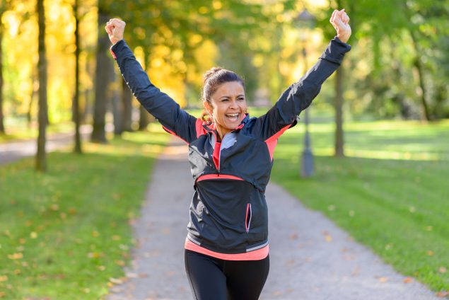 Mujer con ropa deportiva y con los brazos en alto haciendo un gesto de victoria mientras corre al aire libre.