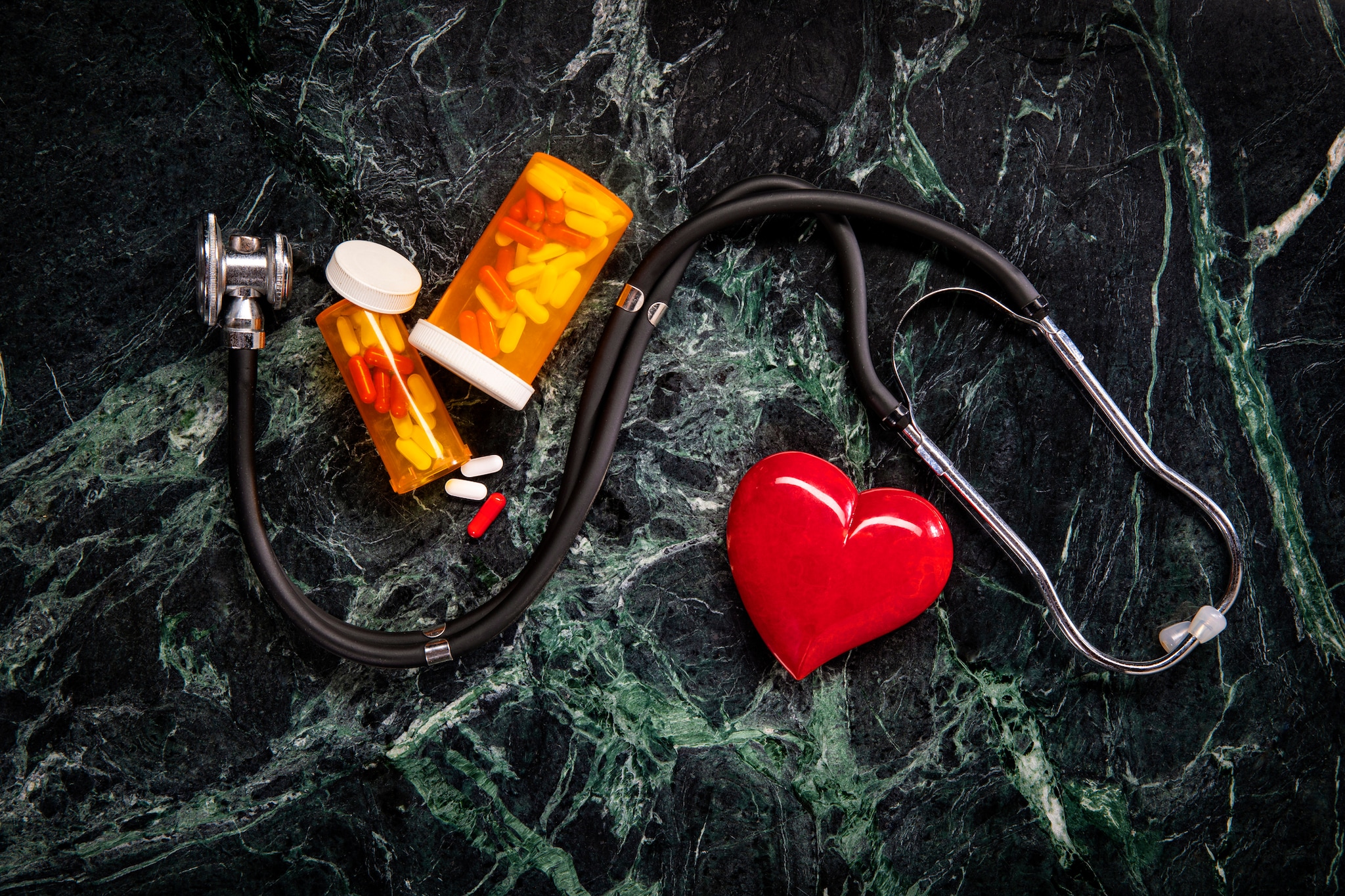 Estetoscopio, frascos de medicamentos y corazón de plástico rojo con un fondo de mármol oscuro.