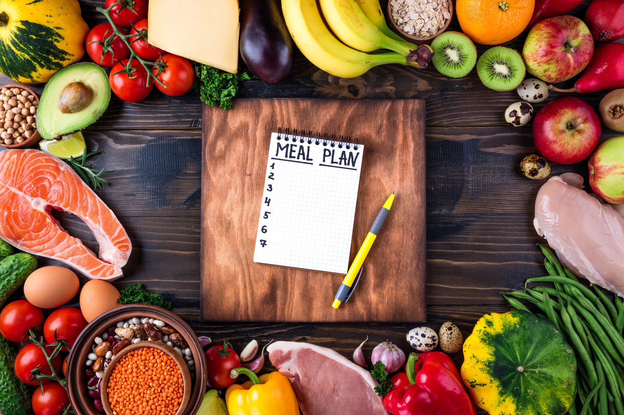 Top view of a meal plan notebook in the center of a wooden table surrounded by fresh vegetables, fruits, meat and fish.