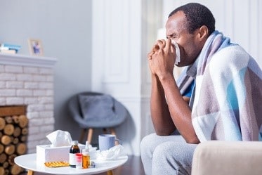 Hombre estornudando en su casa. Se ven medicamentos sobre la mesa.