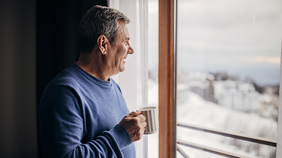 Man holding a mug looking out a window at a winter landscape