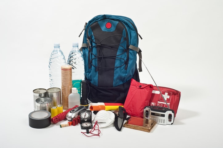 Suministros de emergencia que incluyen una mochila, un kit de primeros auxilios, alimentos enlatados y agua embotellada sobre una mesa.