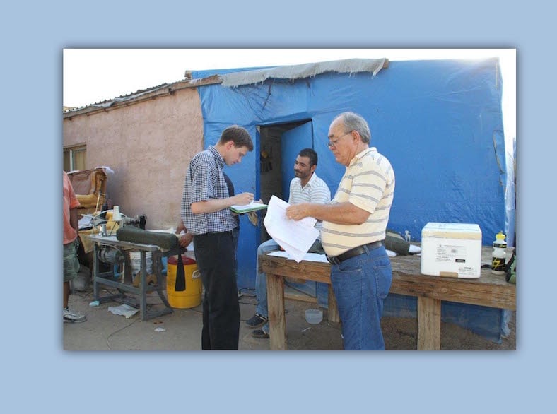 Disease expert from CDC holds clipboard and talks to a resident sitting on a table in front of his house