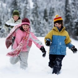Three young children running through the snow wearing winter clothing