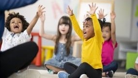 Young children sitting on floor and raising hands in classroom.