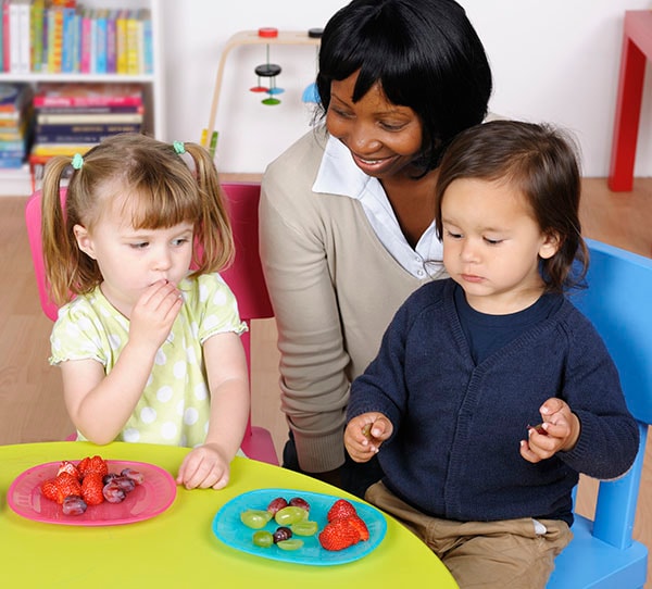Early care worker watch children sit and eat fruit at a table