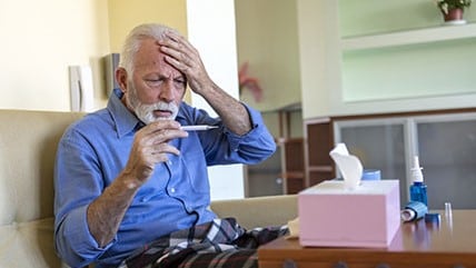 Older man looking at a thermometer with a hand to his forehead.