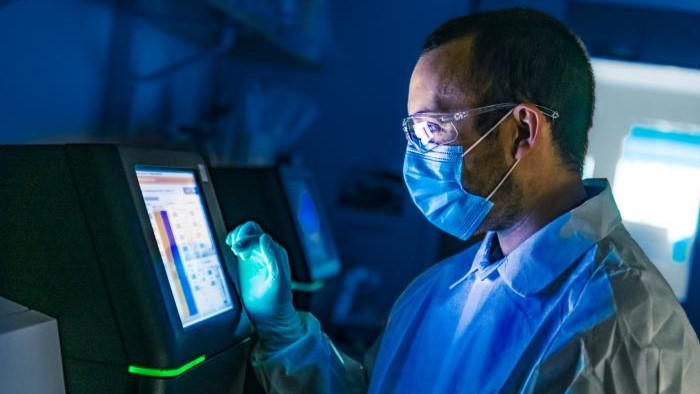 An older adult with protective glasses on their eyes, wearing a face mask over their mouth and nose, looking at genetic analysis results on a monitor.