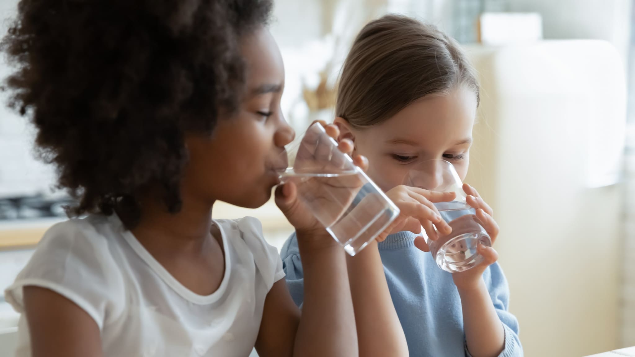 Two children drinking glasses of water next to each other indoors.