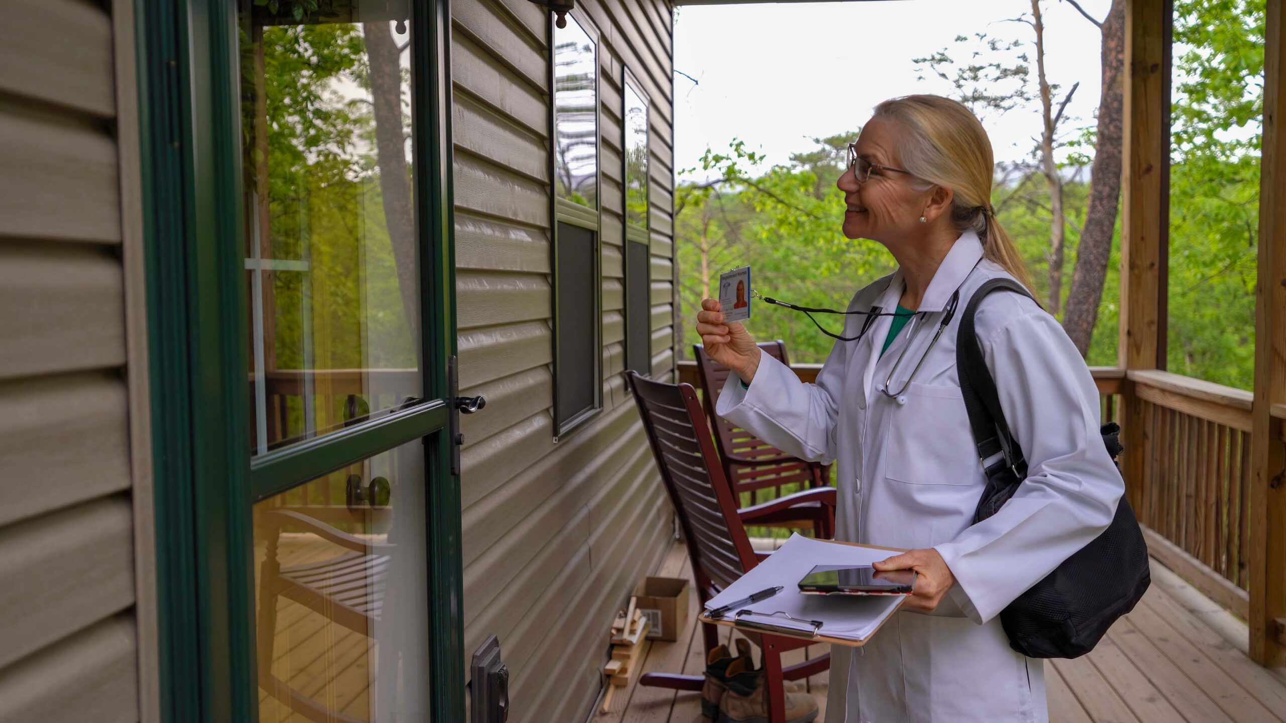 Doctor in lab coat holds up ID badge at a porch door, carrying a clipboard and bag, forest in background.