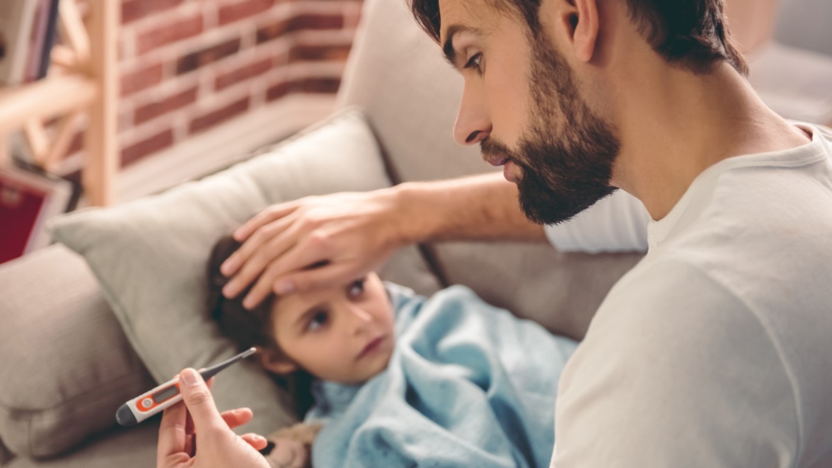 A father takes the temperature of a small child lying on a couch.