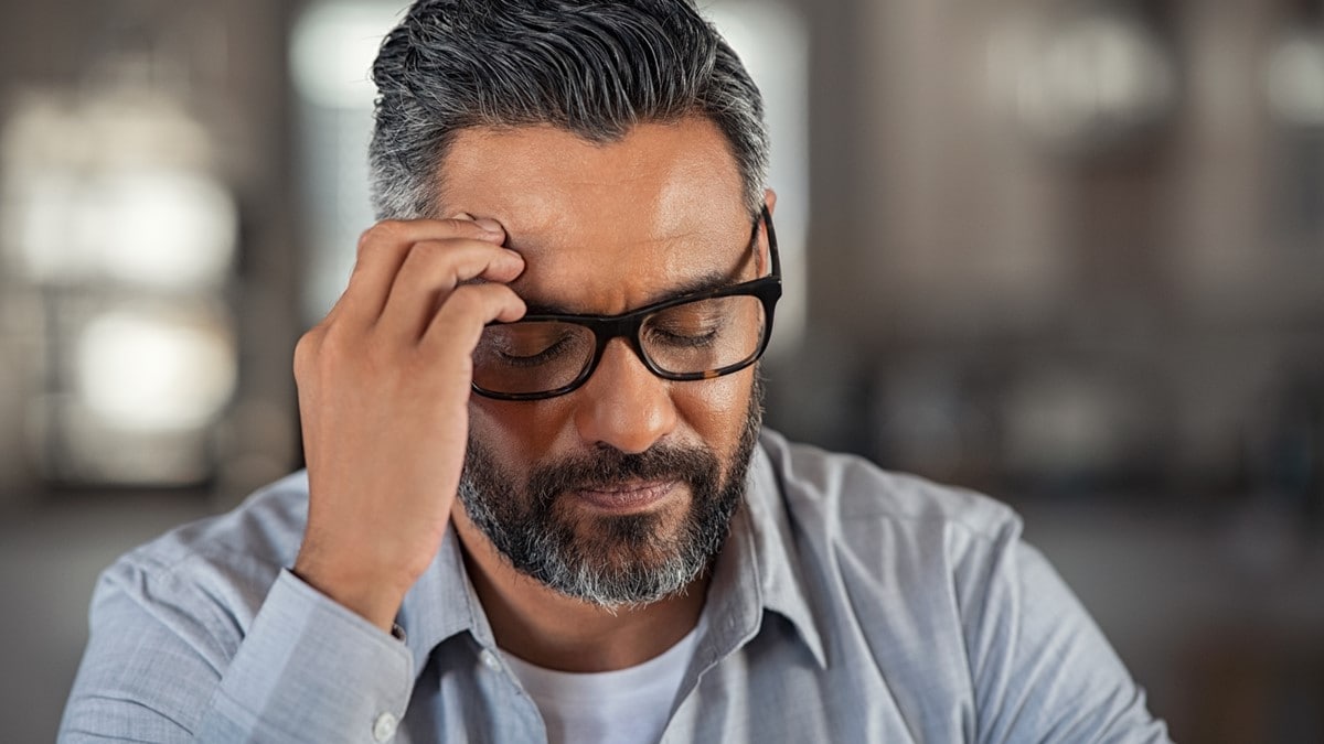 A man wearing glasses puts his hand to his head, as if he is experiencing pain.