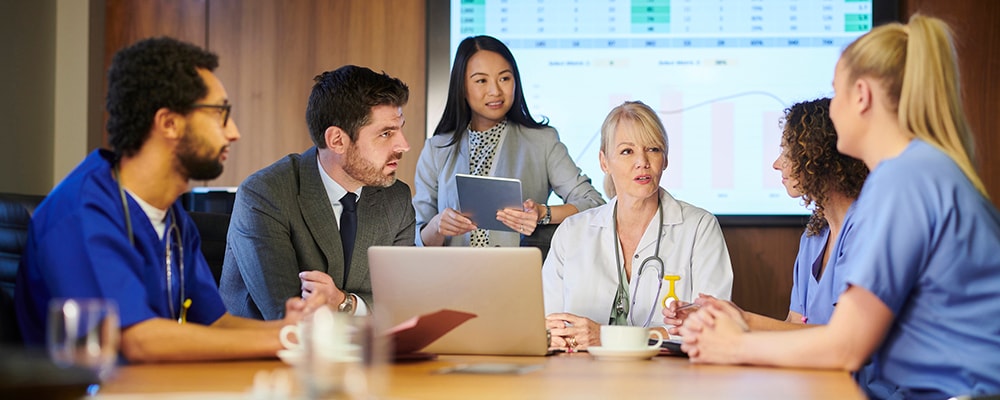 A group of six people are attending a business meeting around a conference table. There is data on the screen behind them. Several are healthcare professionals wearing scrubs or stethoscopes.