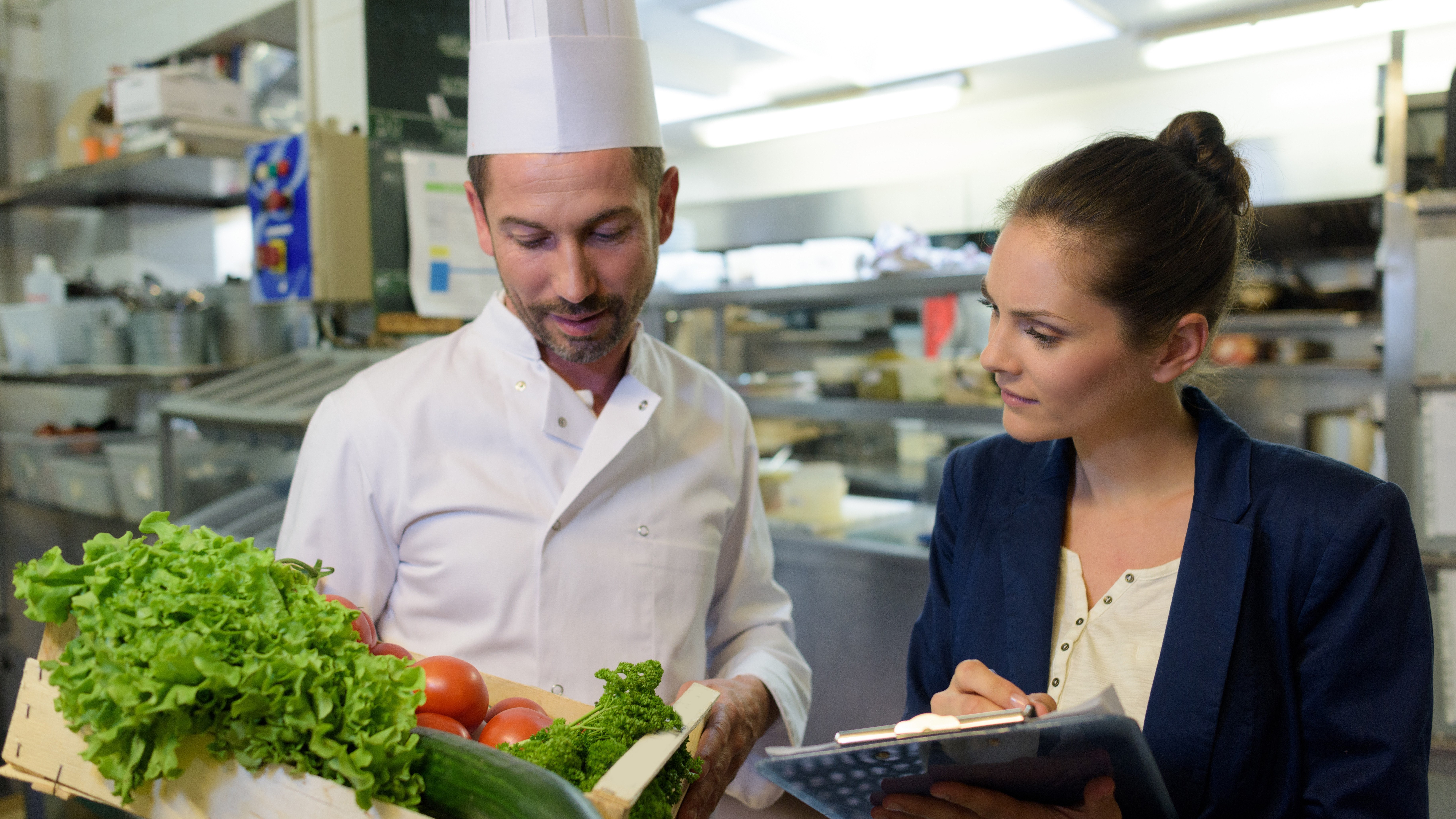 Chef and food inspector in restaurant kitchen in front of box of vegetables