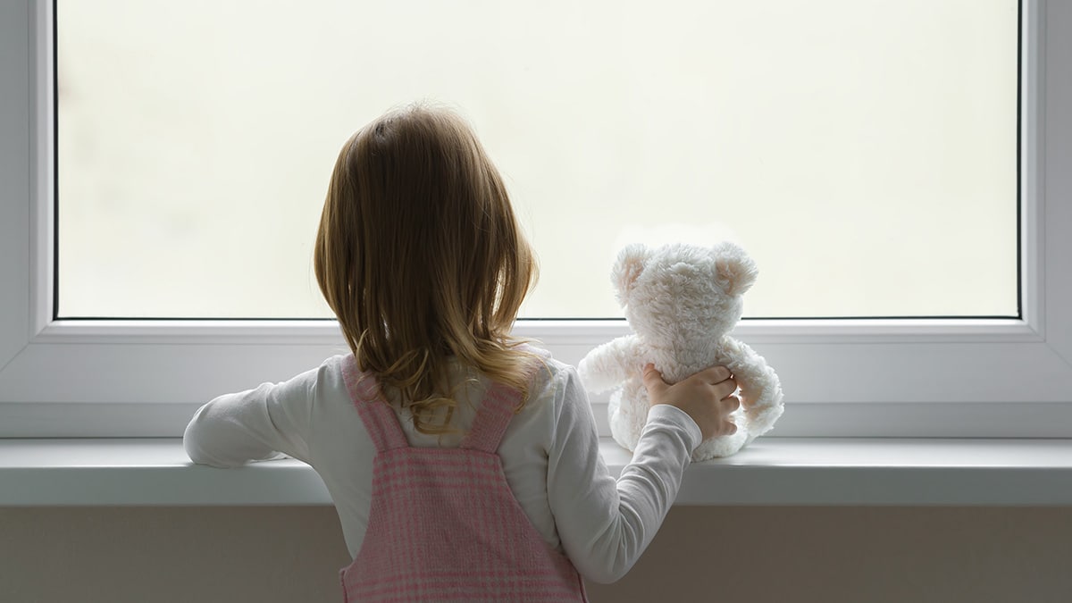 Young girl with teddy bear looks out of window.