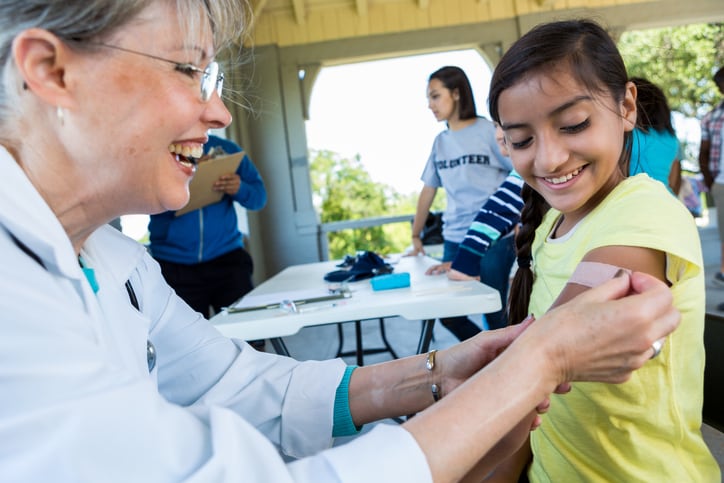 Senior female doctor smiles while putting an adhesive bandage on a preteen girl's arm following an immunization. The doctor is volunteering at an outdoor health fair.