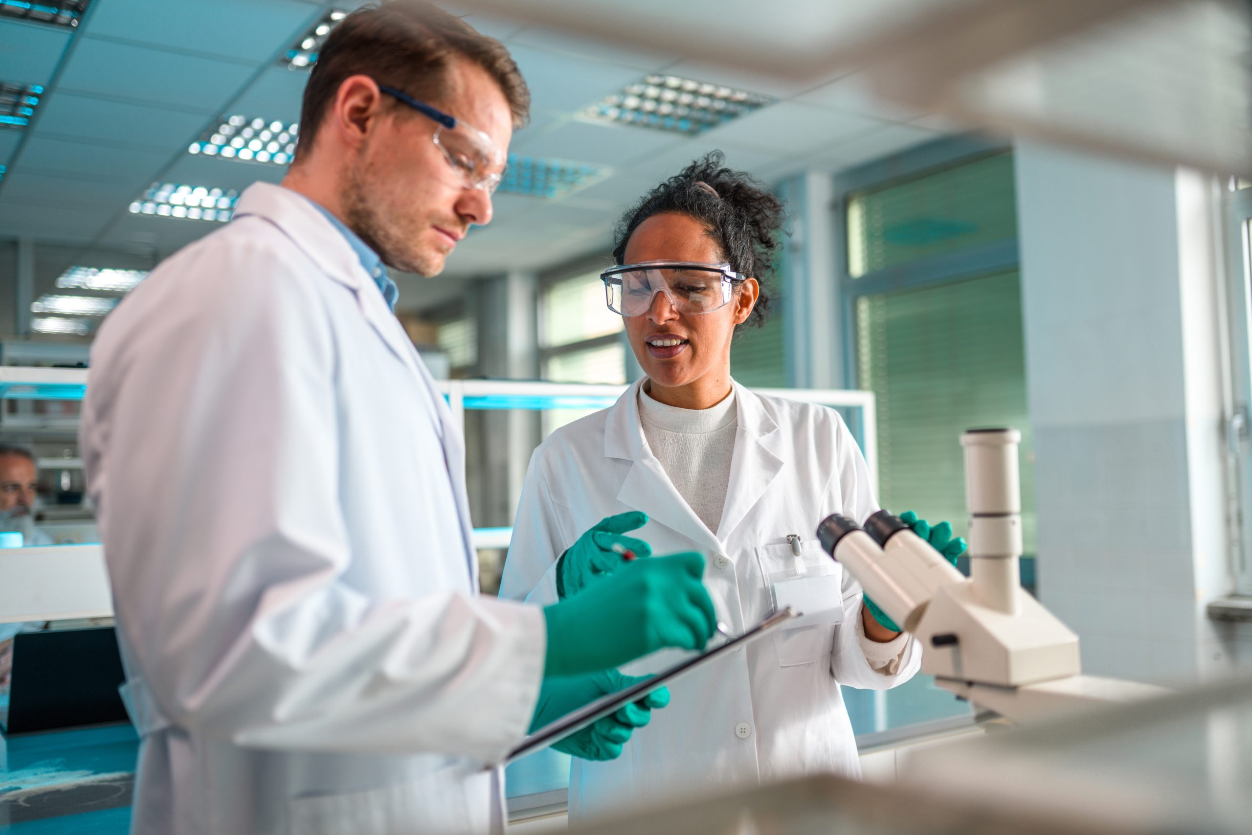 Female and male researchers working in a medical laboratory wearing white coats, surgical gloves, and glasses.