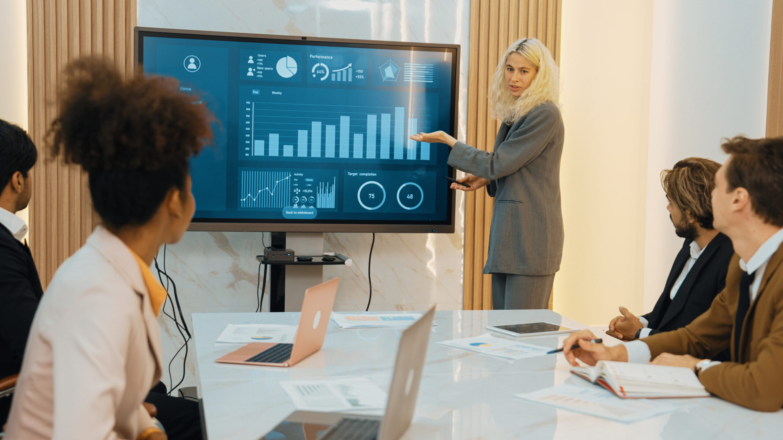 A business professional stands in front of a diverse group of colleagues and presents their work to the group.