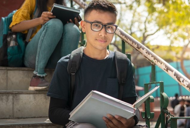 A teen boy sits on outside stairs holding an open book.