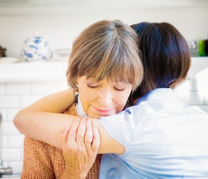 An older Caucasian woman being hugged by another woman in a kitchen. The older Caucasian woman’s face is shown. Her eyes are closed and she has a calm look on her face. 