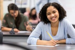 Teen girl smiles as she sits in a classroom, writing on a piece of paper.