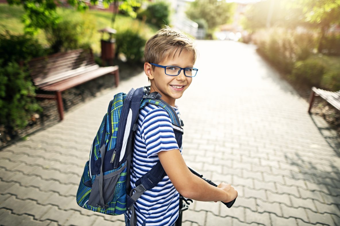 Elementary school aged boy with glasses and a backpack walks down a path to school.