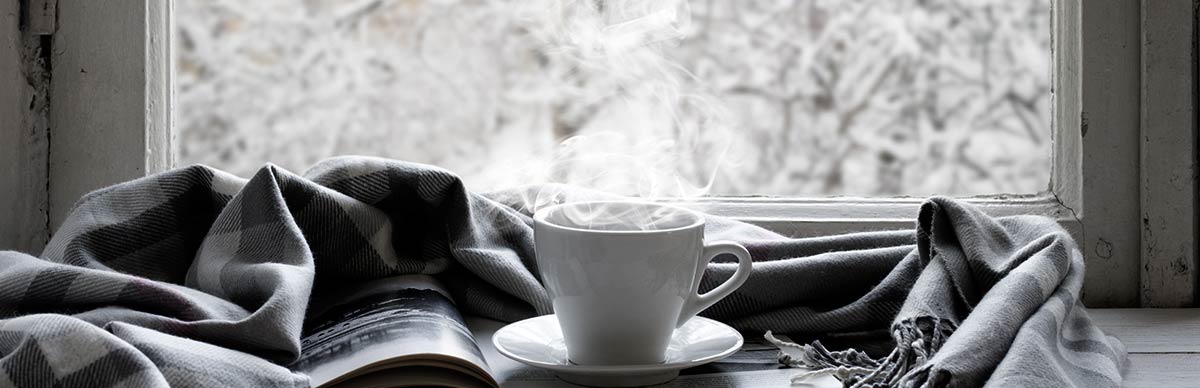 Un libro, una bufanda gris y blanca y una taza blanca llena de una bebida caliente y humeante se colocan en una mesa frente a una ventana que muestra un paisaje nevado afuera.