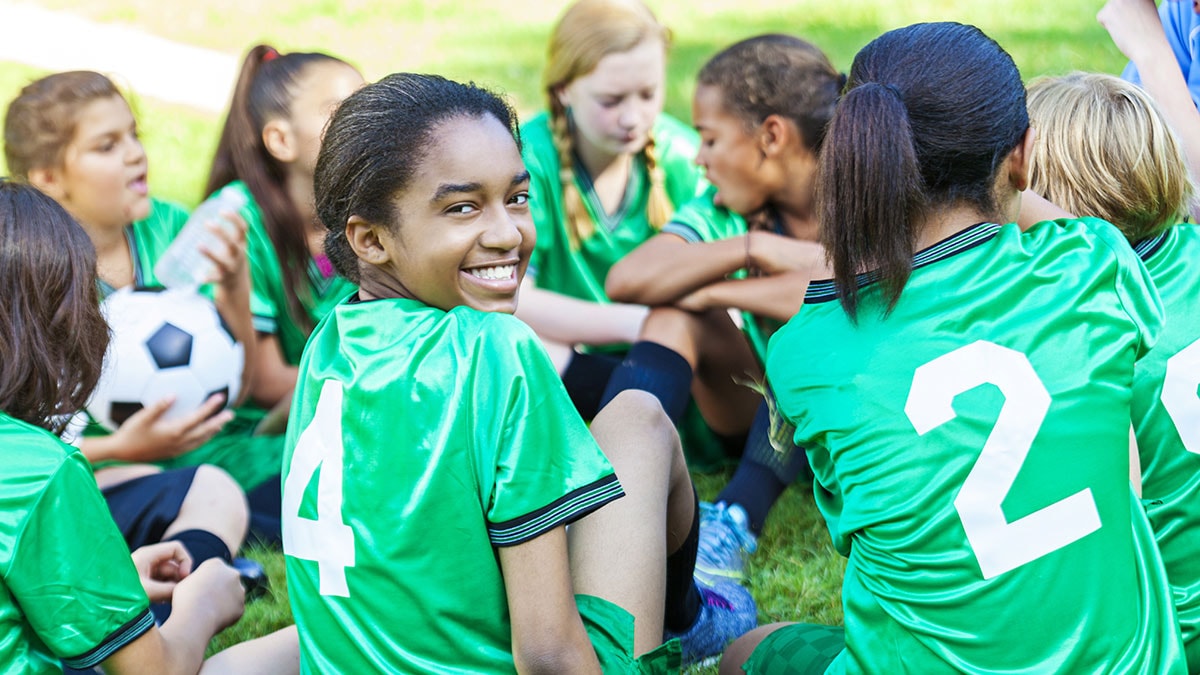 Group of teenage girl athletes sitting on the grass wearing green soccer jerseys while one is holding a soccer ball.