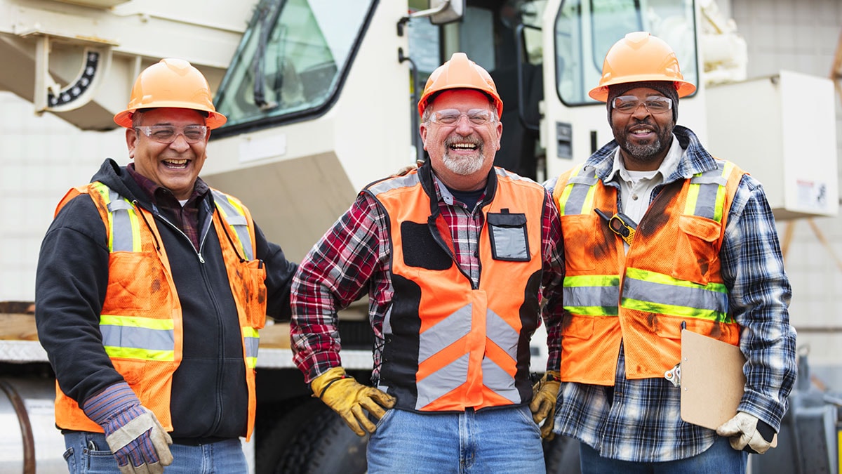 Three smiling construction workers standing side-by-side in front of equipment.