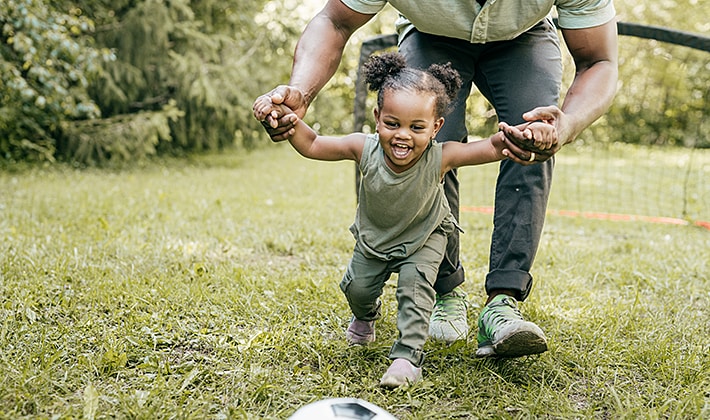 Caregiver helping a toddler run towards a soccer ball.