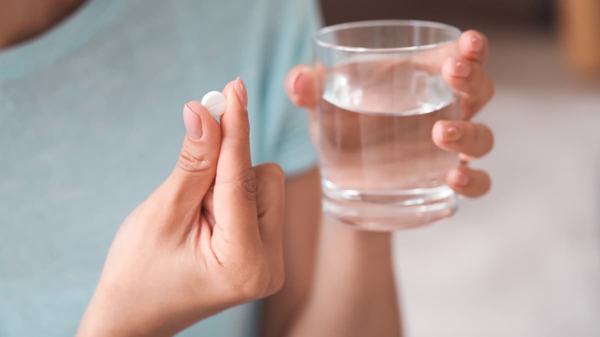 Focused view of a small, white vitamin tablet and glass of water in a person's hand