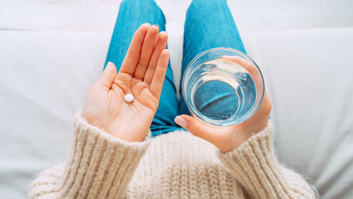 Point of view of a person sitting on the couch holding a vitamin on one hand and a glass of water in the other hand.