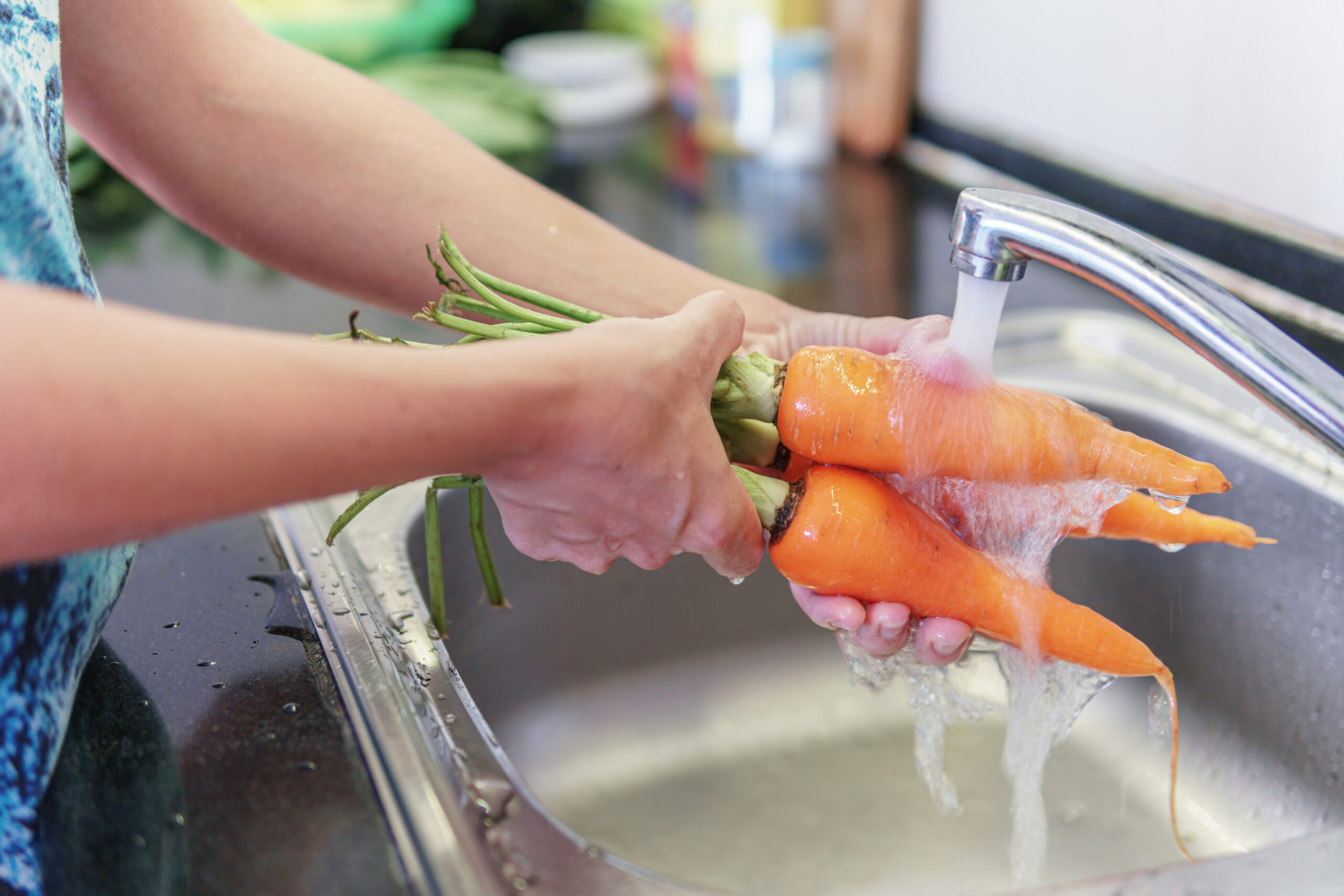 Hand holding carrots and washing them under running water coming out of kitchen sink faucet