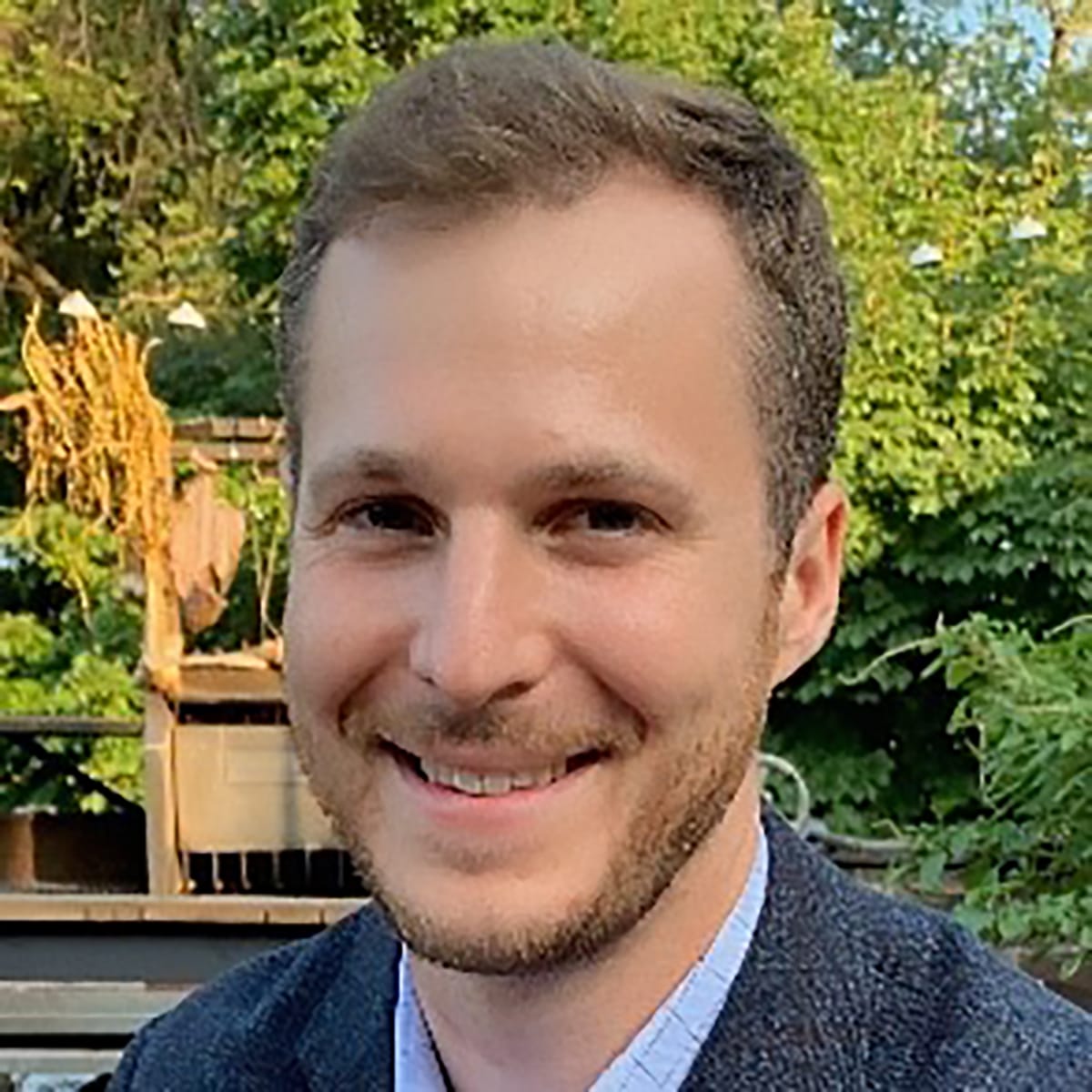 Jason Asher, Ph.D., is the director of the Predict division for the Center of Forecasting and Outbreak Analytics. He has short blond hair, smiling, wearing a grey shirt against a background of green trees.