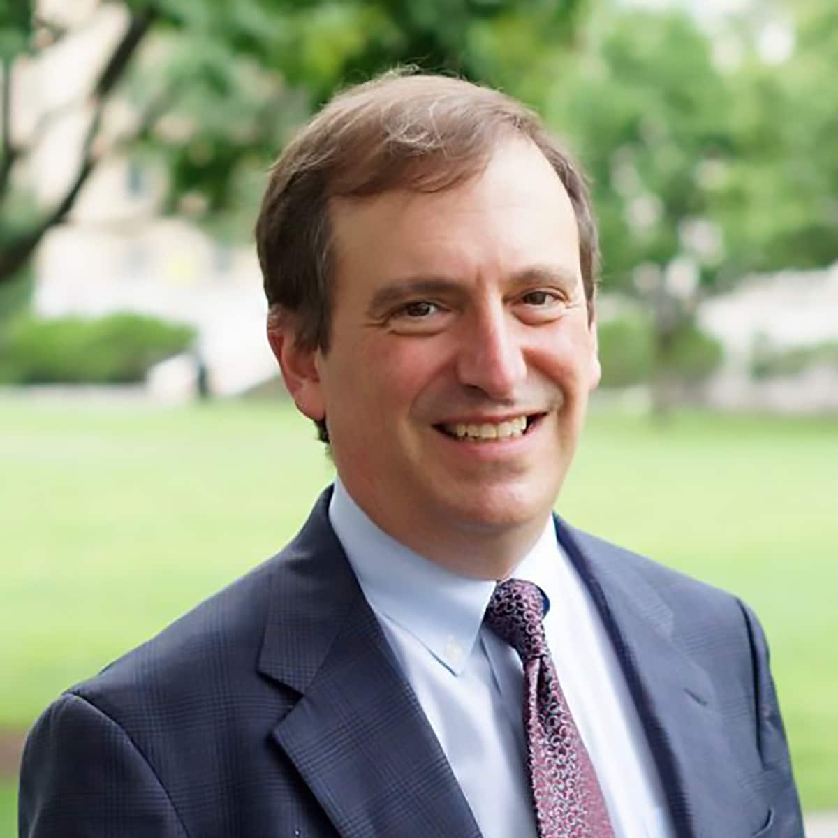 Marc Lipsitch, DPhil, is the senior advisor for the Center of Forecasting and Outbreak Analytics. He has short brown hair, smiling, wearing a grey suit, against a background of green grass and trees.