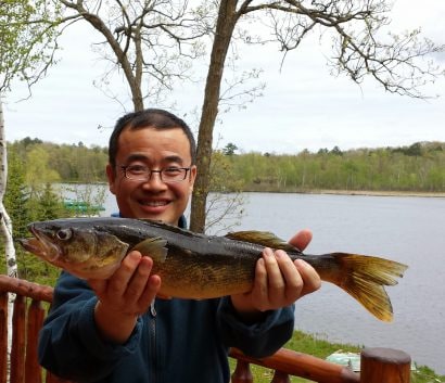 Smiling man holding a fish with both hands next to a lake.