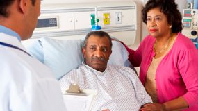  a husband laying in a hospital bed and listening with his wife to the doctor