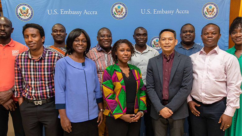 A group of 11 people pose in front of a blue U.S. Embassy Accra banner.