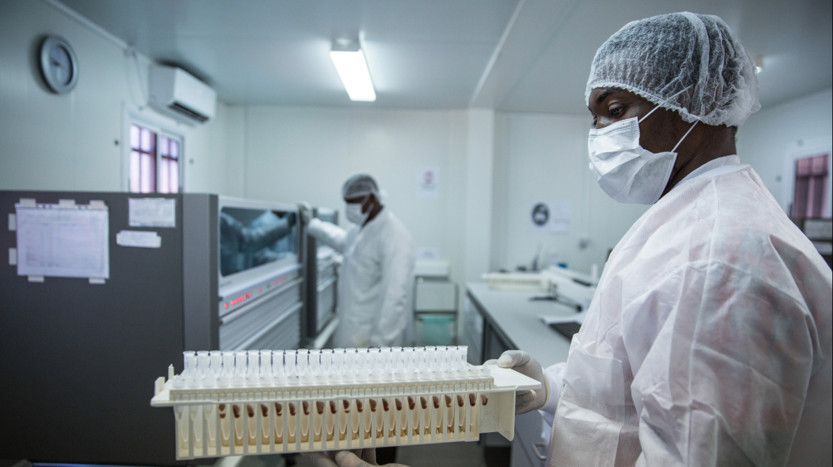 Lab technician in protective gear holding a tray of sample vials, with a colleague working at lab equipment in the background.
