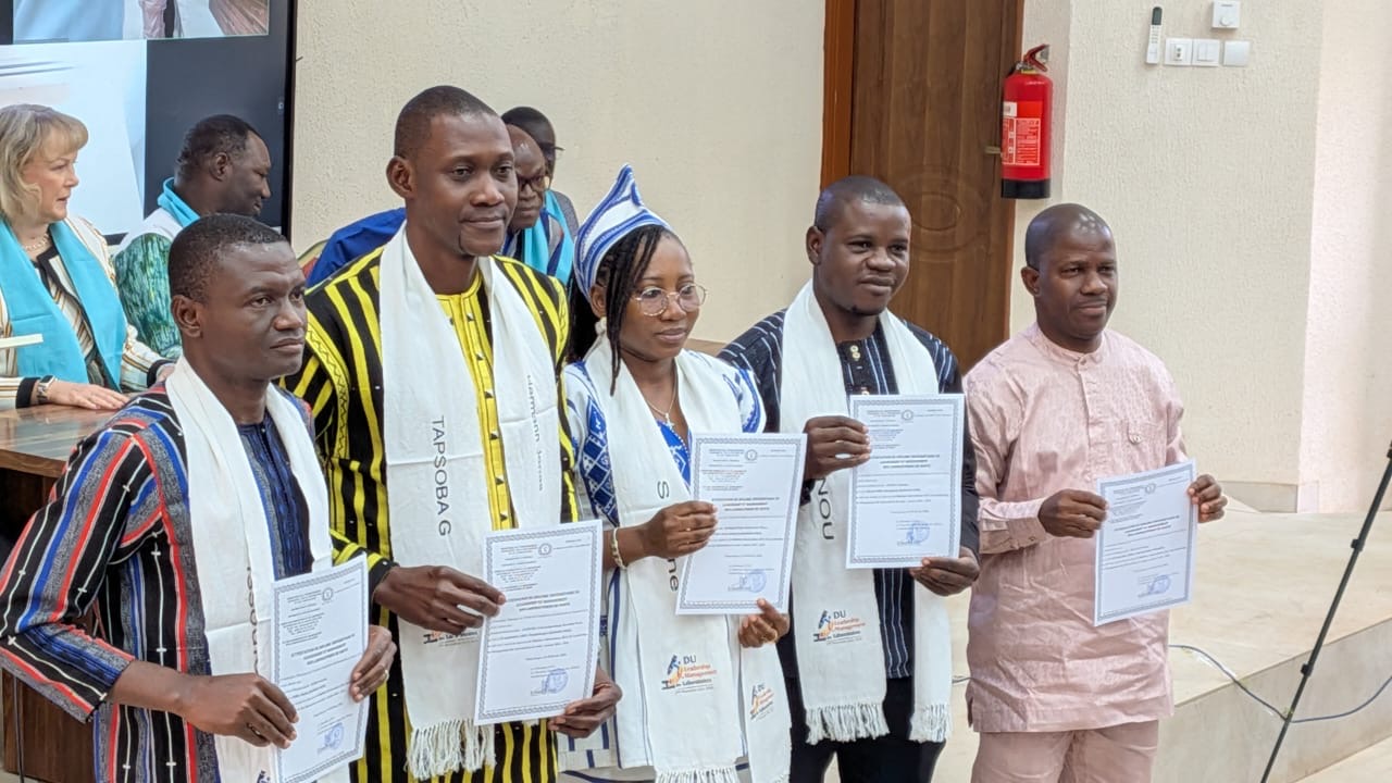 Five people pose with certificates at an indoor ceremony, with four wearing ceremonial scarves.