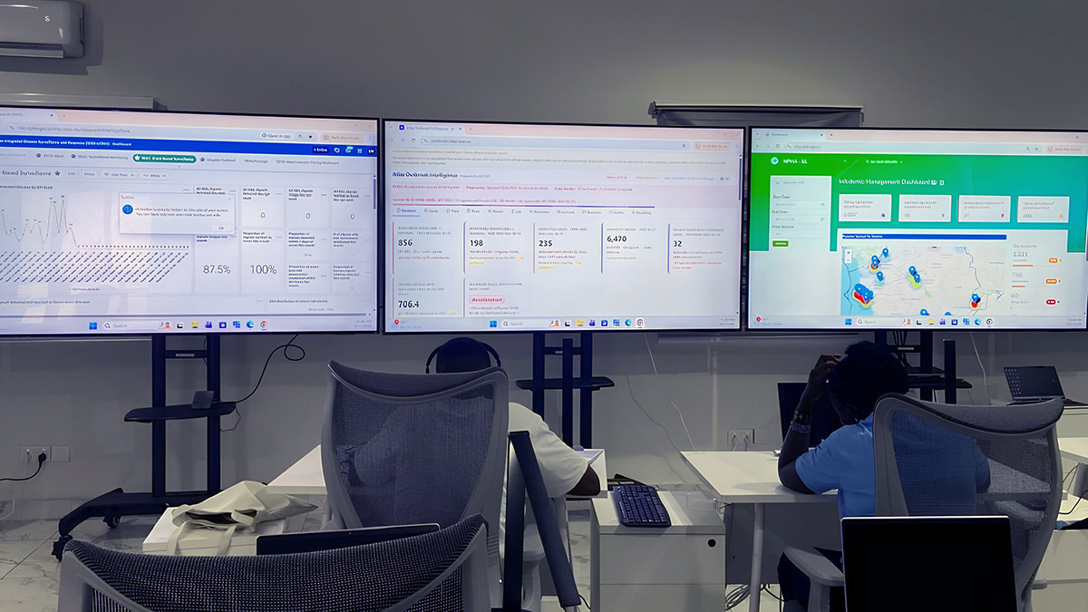 Two staff members monitor three large screens displaying emergency operations dashboards and a map.