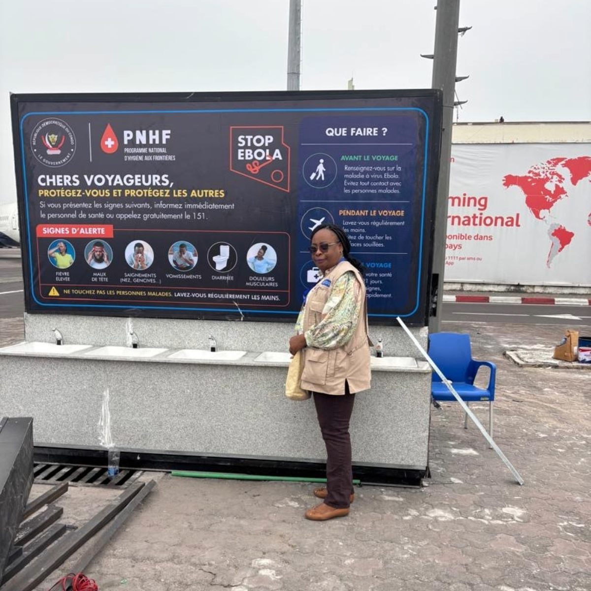 Woman stands beside an Ebola prevention sign and handwashing station for travelers.