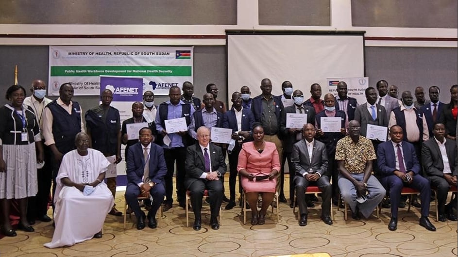 A photo of over 30 people standing or sitting closely together for a group photo. Some people are holding up certificates. A sign behind them reads, Ministry of Health, Republic of South Sudan, Public Health Workforce Development for National Health Security.