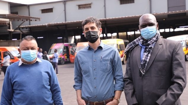 Khankhara, Hines, and Mvula pose for a photo outside a bus depot. All three individuals are wearing face masks.